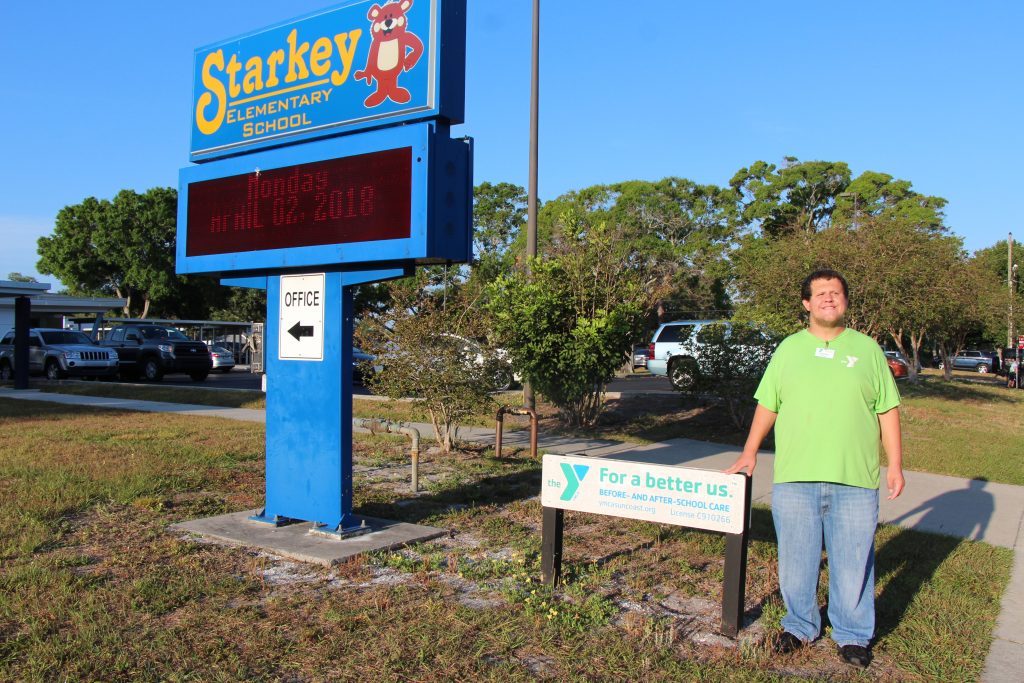 Justin smiling standing next to a YMCA sign outside of an Elementary School