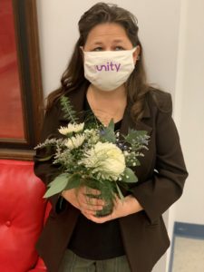 Jessica is wearing a brown suit jacket and grey dress slacks. She hold a bouquet of white and green flowers wearing a face mask as she smiles at the camera. 