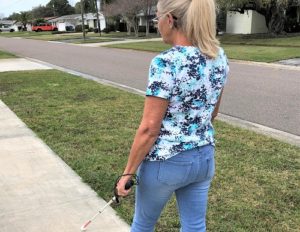 Susan walks along a neighborhood street holding a white cane. she is wearing a blue shirt and blue jeans and is walking away from the camera.