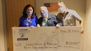 Judge Joe Donahey holds up a a large check for $50,000 that will be donated to lighthouse of Pinellas. He looks off to the right of the camera, wearing a face mask and smiling. His son stands to the left of him wearing a face mask smiling at the camera. 
